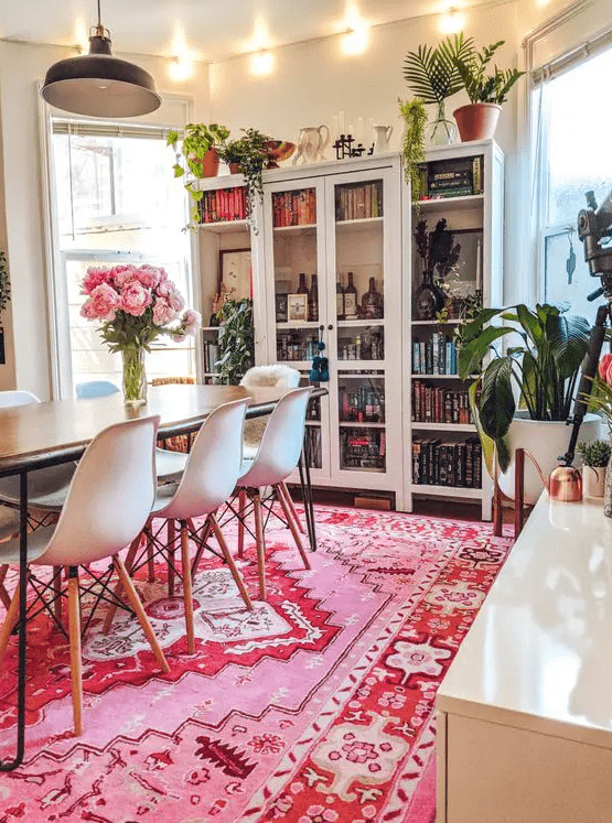 a modern dining room done in neutrals, with a large bookcase, a table and white chairs, lots of potted greenery and a bold pink rug to add color