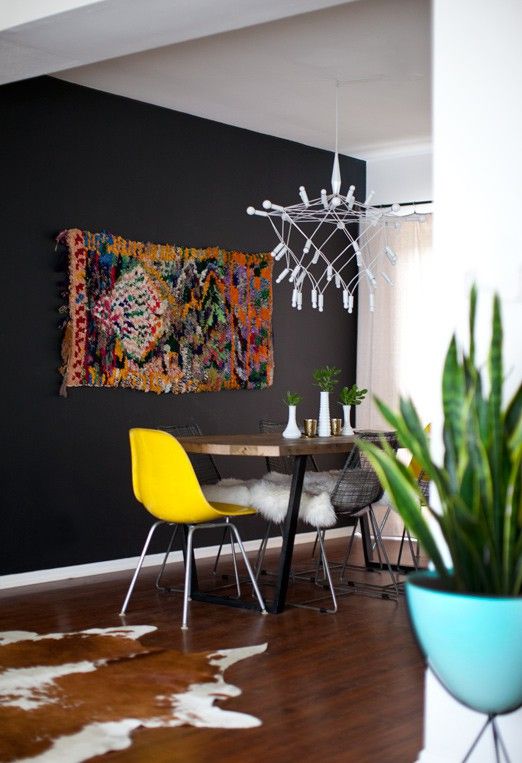 a modern dining room with a black accent wall, a bright boho rug, a stained table and mismatching chairs, a whimsical modern chandelier