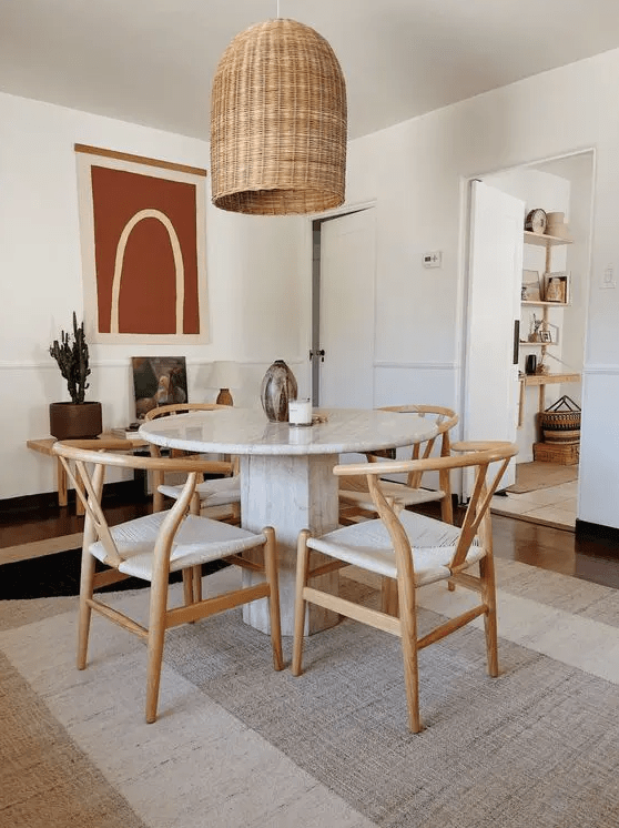 a modern dining room with layered rugs including a jute one, a round table, woven chairs, a woven pendant lamp, a bench with decor is a chic space