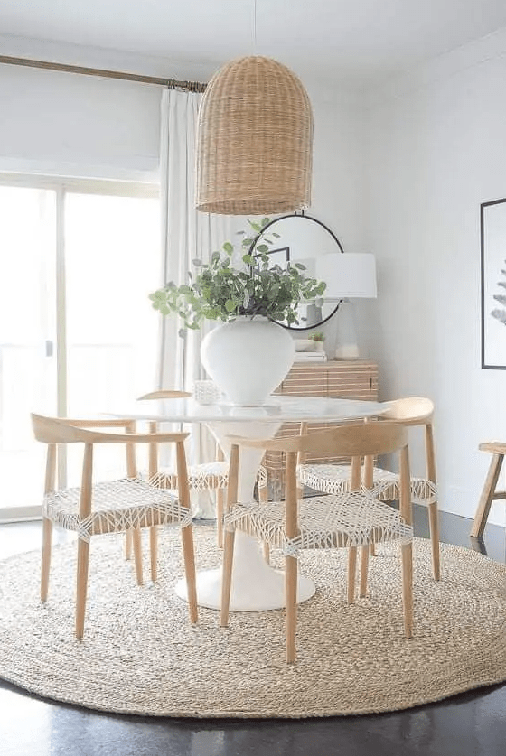a modern dining space with a white table, woven chairs, a jute rug and a woven pendant lamp plus some chic decor