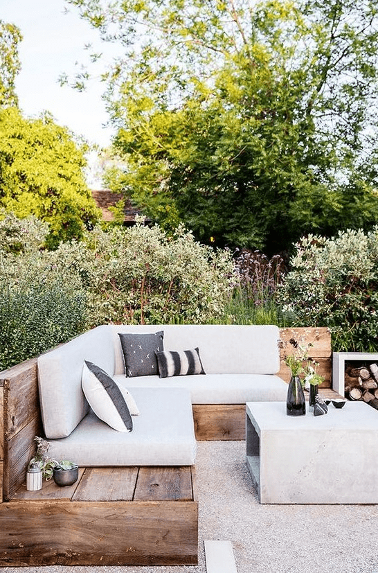 A modern farmhouse outdoor living room with a large built in corner sofa, a concrete coffee table, printed pillows, a firewood storage unit