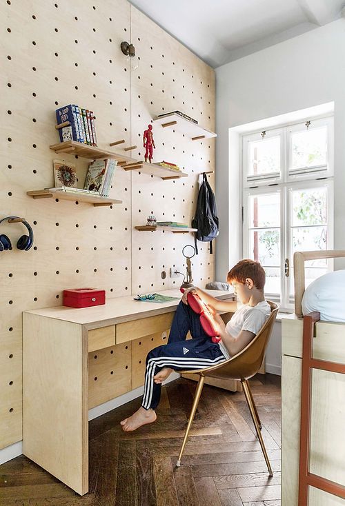 a modern kids' room with a pegboard wall that holds shelves and various stuff, with a stained desk and a neutral chair