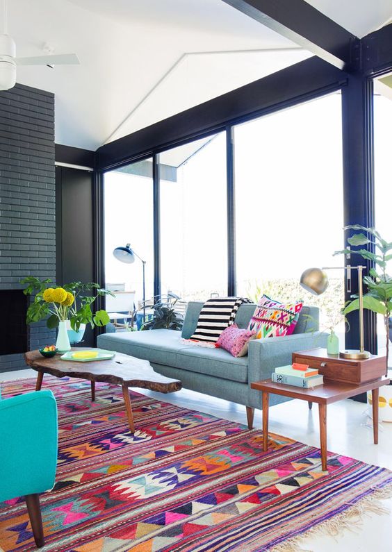 a modern living room with a fireplace, a grey sofa with colorful pillows, a bright boho rug, a turquoise chair and greenery