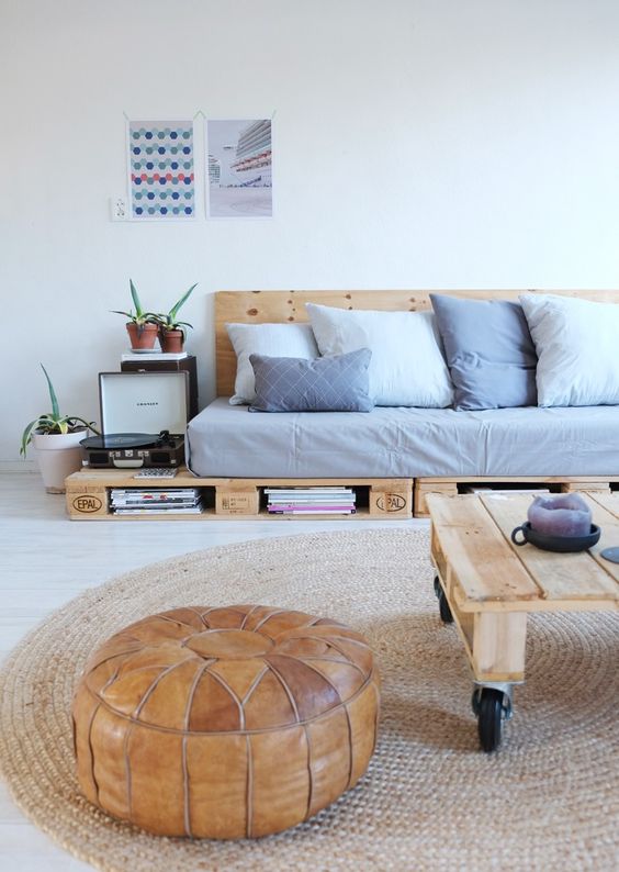 a modern living room with a pallet sofa and pillows, books inside, a pallet coffee table on castersm a leather pouf and some greenery