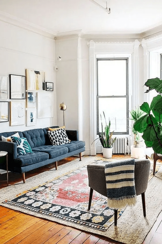 a modern living room with layered rugs, a jute and boho rug, a navy sofa with pillows, a grey chair, a gallery wall and potted plants
