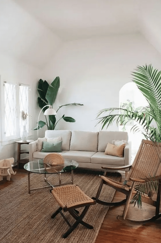 a modern neutral living room with a neutral sofa, a woven rocker and a footrest, a jute rug, a glass coffee table and potted plants is a chic space