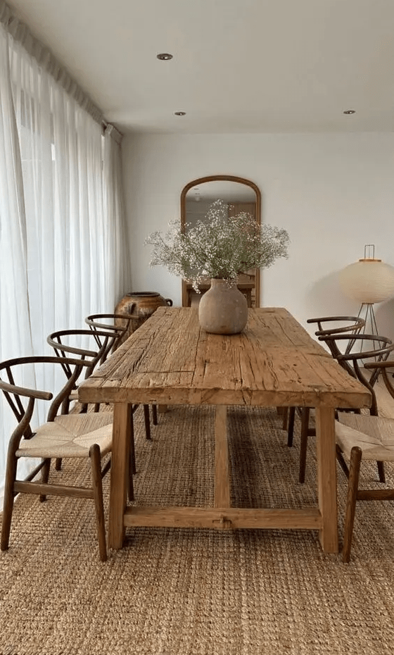 a modern rustic dining room with a jute rug, a rough wood table and woven chairs, a mirror, a lamp and some blooms in a vase