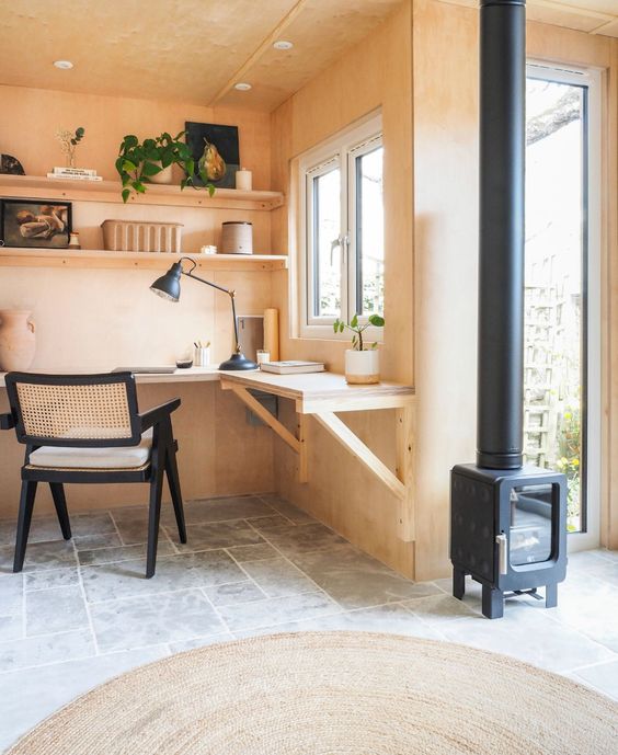 A modern shed home office with MDF walls, a wall mounted corner desk, a cane chair, a hearth and some decor and potted plants