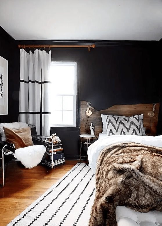 a moody eclectic bedroom with black walls, a bed with a living edge headboard, neutral and printed bedding, a leather chair, a book stack and printed textiles