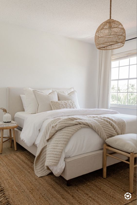 a neutral bedroom with a jute rug, a neutral bed with neutral bedding, wooden chairs, a woven pendant lamp and some decor