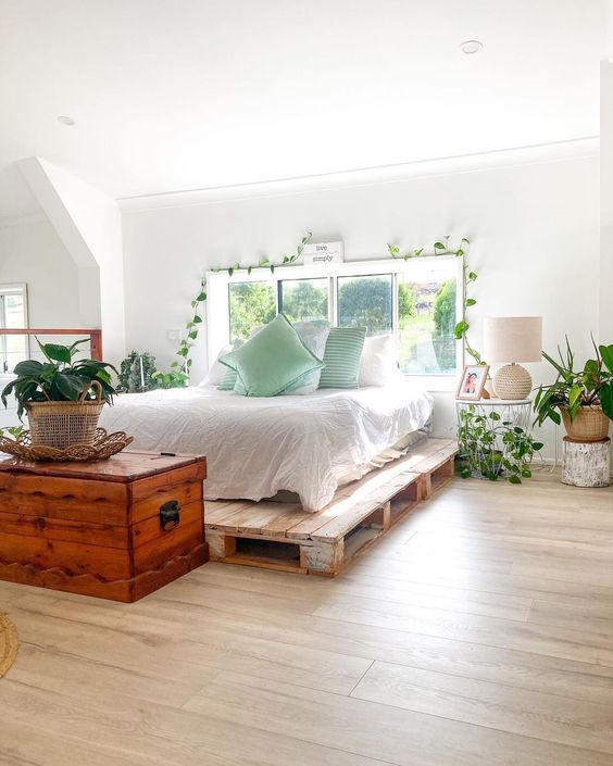 a neutral bedroom with a pallet bed and neutral bedding, a chest, some potted plants and greenery plus a table lamp