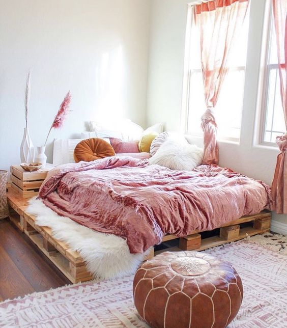 a neutral bedroom with a pallet bed and pink bedding, pink curtains, a leather pouf, some pillows and pampas grass