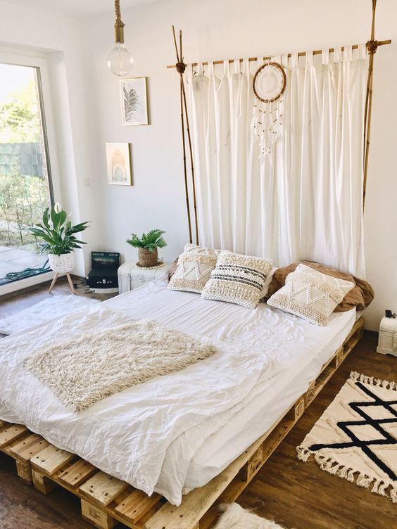 a neutral boho bedroom with a floor to ceiling window, a pallet bed with neutral bedding, some rugs and a curtain headboard plus potted greenery