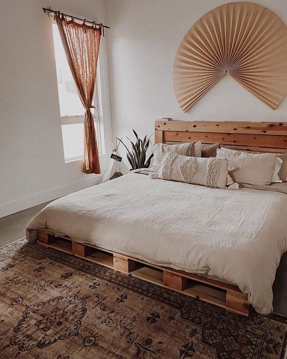 a neutral boho bedroom with a pallet bed and neutral bedding, a boho rug, a fan and a rust curtain is a lovely space