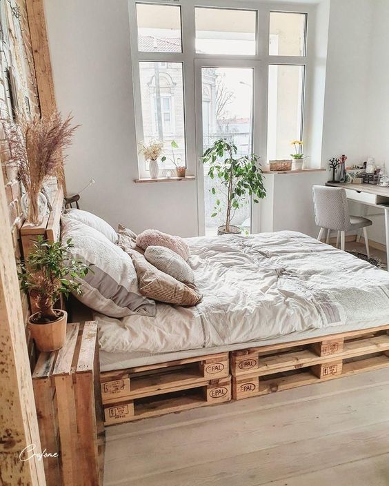 a neutral boho bedroom with a pallet bed and neutral bedding, a desk and a grey chair, potted plants, boho decor and lots of pillows