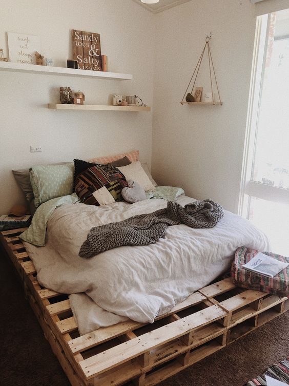 a neutral boho bedroom with a pallet bed and neutral bedding, shelves with decor and some bright pillows