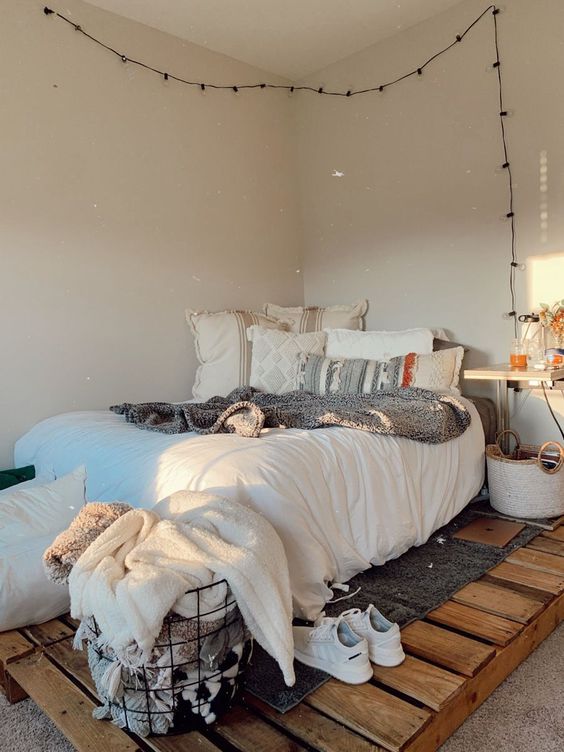 a neutral boho bedroom with a pallet bed, neutral bedding, a wall-mounted nightstand, lights and a wire basket with blankets