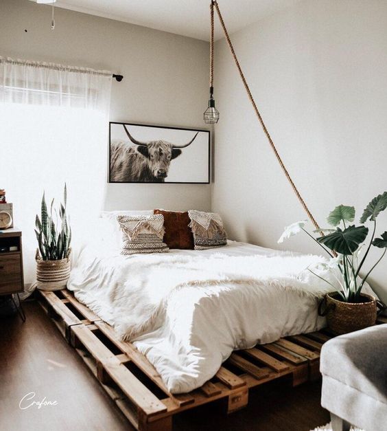 a neutral boho bedroom with a pallet bed with neutral bedding, a stained storage unit, potted plants, a pendant lamp and some art