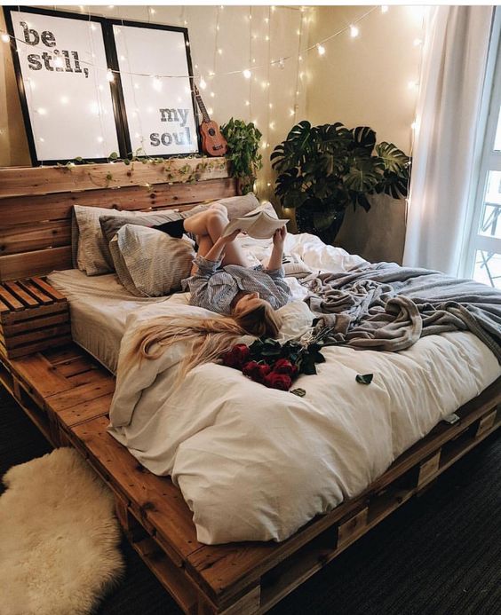 a neutral boho bedroom with a stained pallet bed, neutral bedding, some rugs, lights, decor and a statement plant in the corner