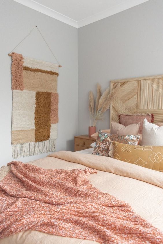 a neutral boho bedroom with grey walls, a bed with pastel pink bedding, a boho rug on the wall, some pampas grass