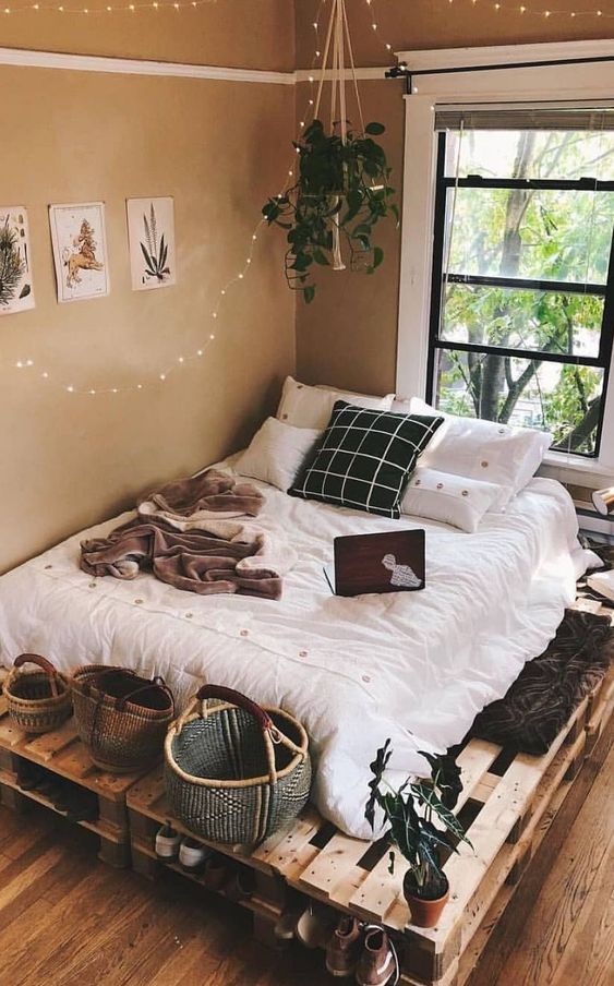 a neutral boho bedroom with tan walls, a pallet bed with neutral bedding, baskets and lights plus greenery
