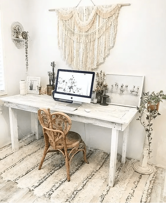 a neutral boho home office with a whitewashed desk, a unique rattan chair, a macrame hanging, potted plants and artworks