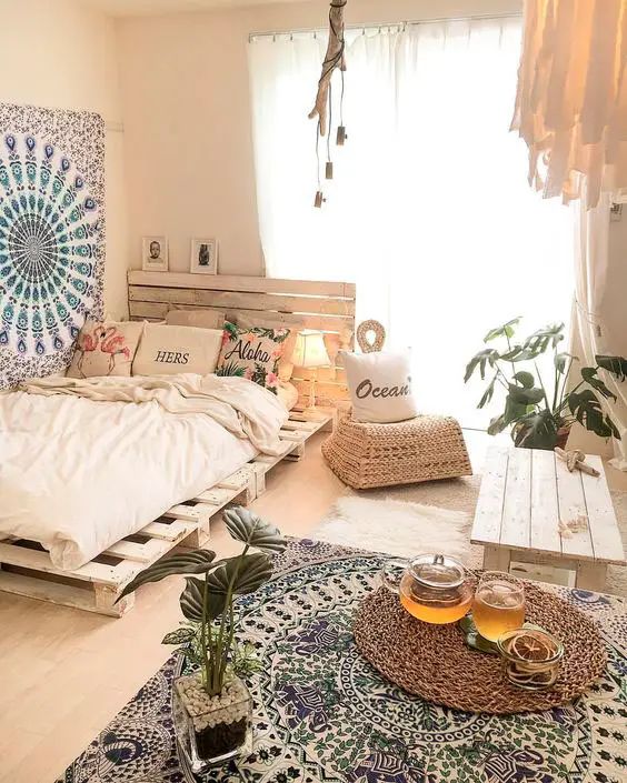 a neutral eclectic bedroom with a whitewashed pallet bed, a boho rug on the wall and floor, a table, a wicker chair and some bulbs
