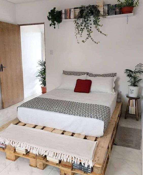 a neutral modern guest bedroom with a pallet bed and neutral bedding, a shelf with books, potted plants and blankets