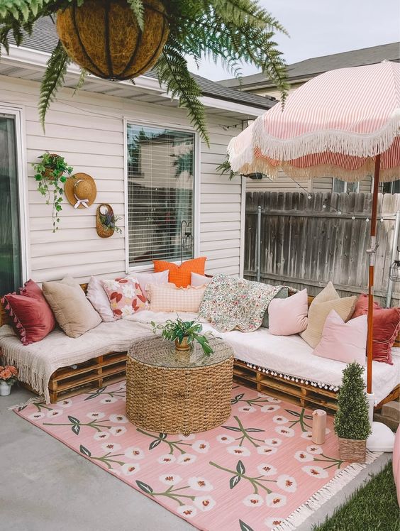 a pink boho terrace with a pallet corner sofa and lots of pillows, a jute table, some greenery, a pink boho rug and a striped umbrella