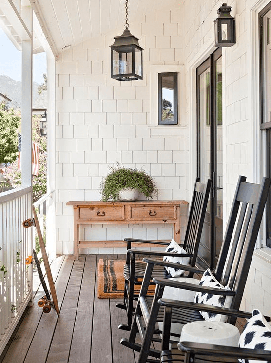 a pretty and simple farmhouse porch with a vintage console table, black rockers and printed pillows, potted greenery and some lanterns is welcoming