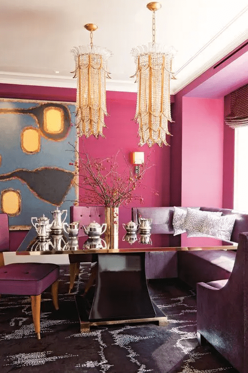 a quirky glam dining space with hot pink walls, a refined black and gold dining table, a built-in purple bench, two crystal chandeliers and an abstract artwork