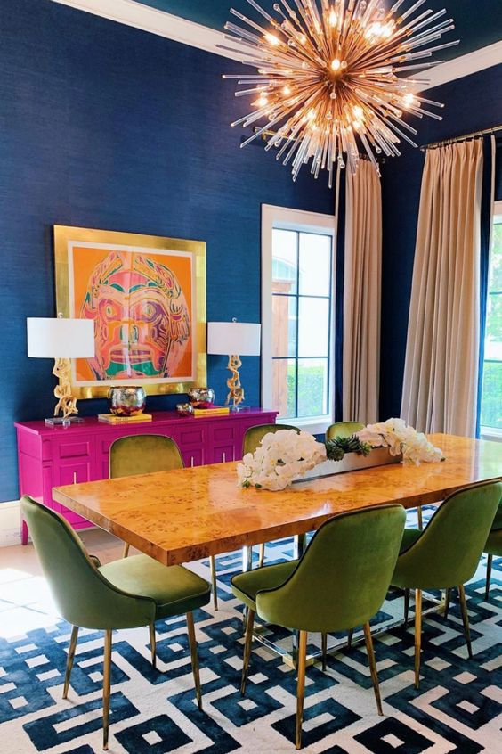 a refined and bold dining room with navy walls, a fuchsia credemza, a table, green chairs, a burst lamp and a bold artwork