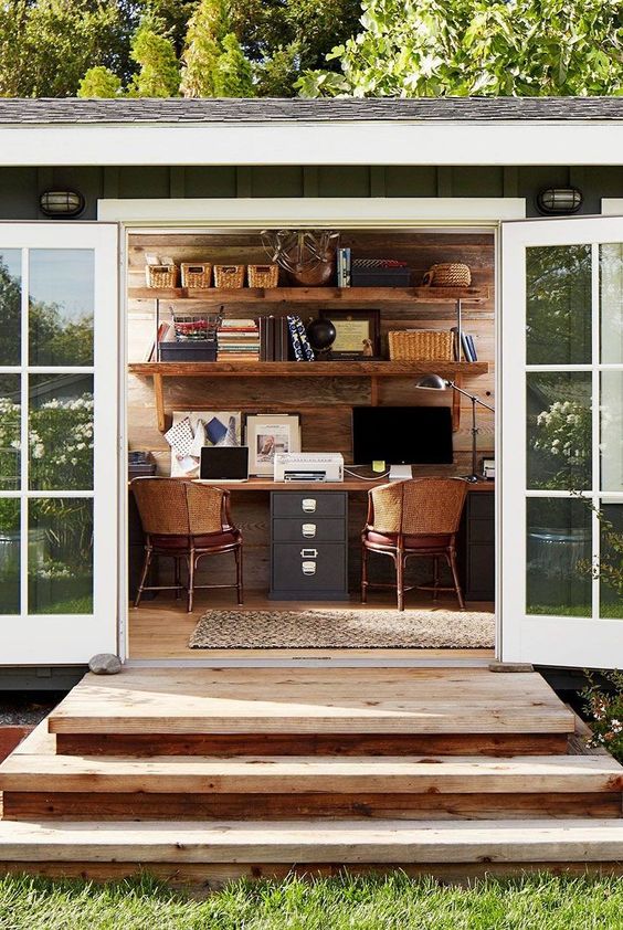 a rustic shed home office with a black double desk, cane chairs, shelves with decor and storage, some lamps and lights