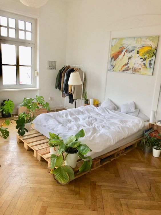 a serene bedroom with a pallet bed and neutral bedding, a makeshift closet, potted plants, an artwork and a lot of natural light