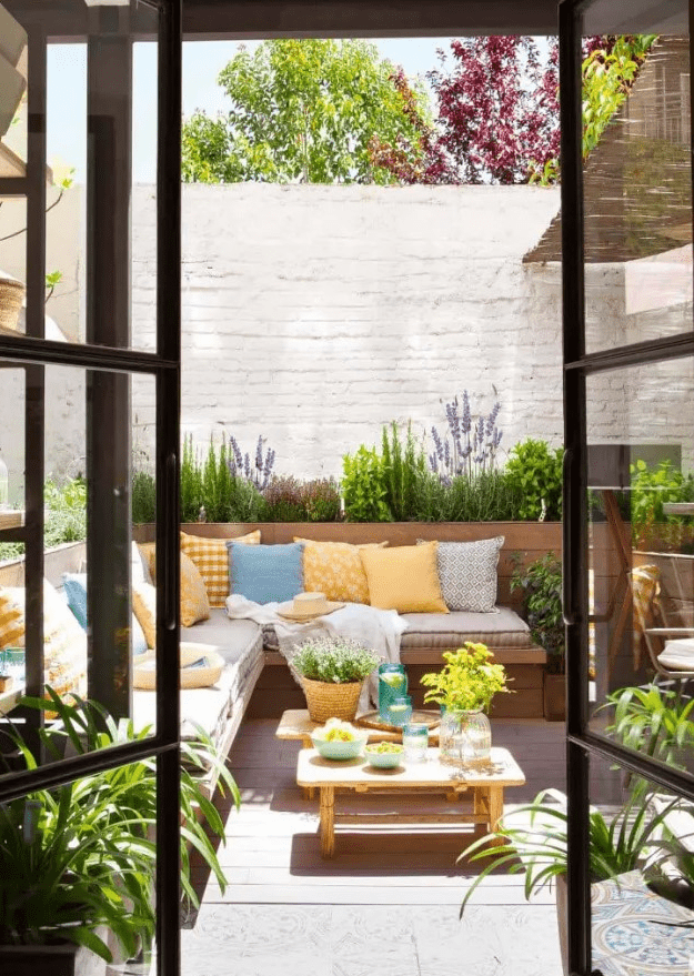 a small and bright patio with a corner sofa with lots of pillows, a couple of benches and blooms and greenery around