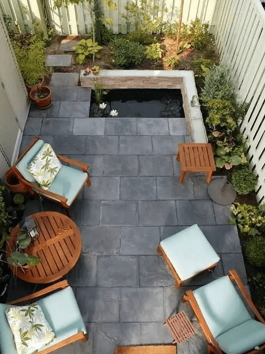 a small and zen-like paved patio with tiles on the ground, a tiny pond, some growing plants and cool garden furniture with blue upholstery