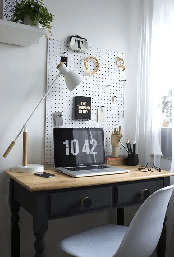 a small eclectic home office nook with a pegboard and hooks, a vintage black desk, a white chair and some stylish decor