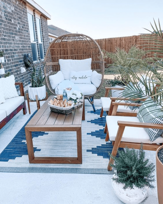 A small patio with white seating furniture, a wooden coffee table, an egg shaped chair, some potted greenery and plants