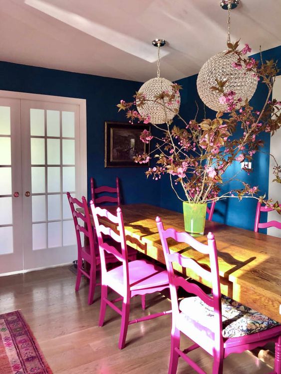 a super bright dining room with navy walls, a long table, fuchsia chairs, pendant lamps and blooming branches is wow