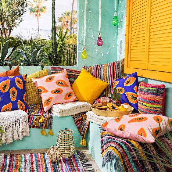 A super bright terrace with aqua walls and yellow shutters, a built in aqua sofa and colorful pillows, bright rugs