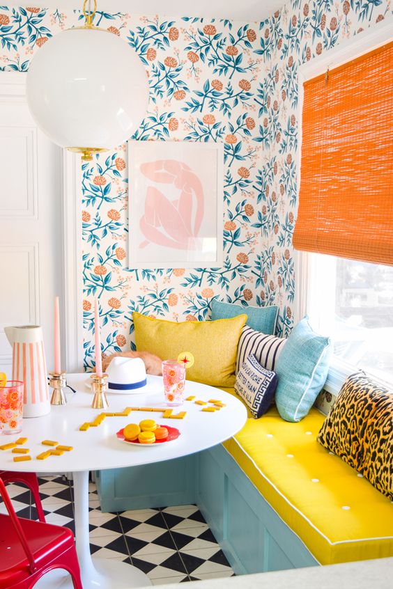 a vibrant dining corner with a turquoise bench and pillows, printed wallpaper, a table and a red chair, orange shades