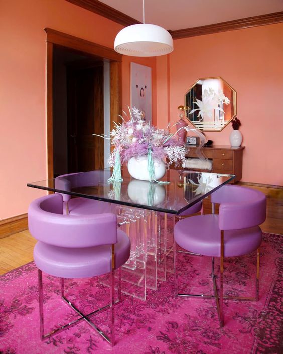 a vibrant dining room with orange walls, a glass table, purple chairs, a fuchsia rug and some chic decor and a mirror on the wall