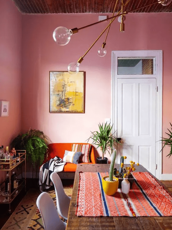 a vibrant dining room with pink walls, an orange loveseat, a dinind table with a colorful runner, some plants and a bar cart
