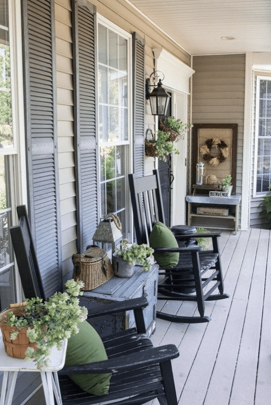 a vintage farmhouse porch with black rockers, a grey chest, potted greenery, a basket, a console and a wreath of wheat