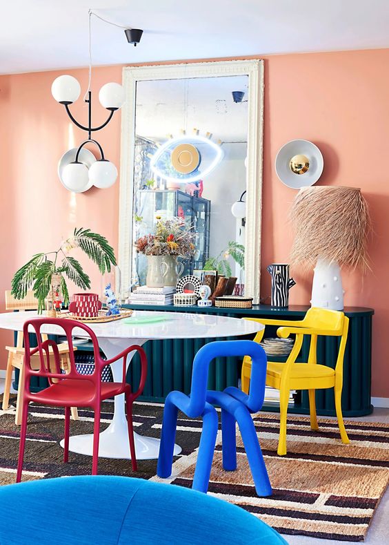 a vivacious dining room with peachy walls, a navy fluted credenza, a table, colorful chairs, a printed rug and some quirky decor