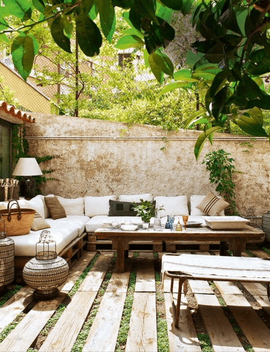 a welcoming Mediterranean summer terrace with pallet and rattan furniture, neutral upholstery and textiles, candle lanterns and greenery
