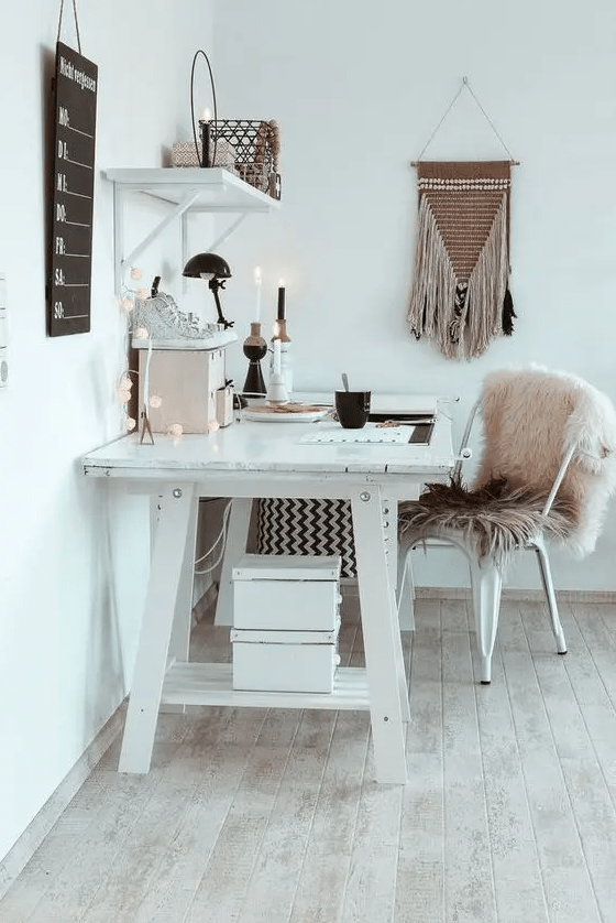 a white boho home office with a large desk, a metal chair with a pink fur cover, candles, lights and a macrame hanging