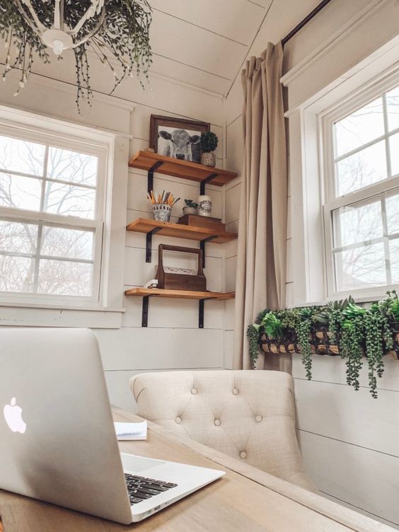 a white farmhouse home office with planked walls, a rustic desk, a creamy chair, shelves with decor and some plants