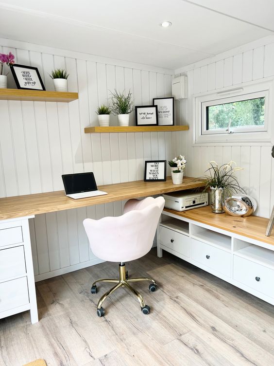 a white home office with planked walls, a desk and a storage unit, shelves with decor and a blush chair with casters