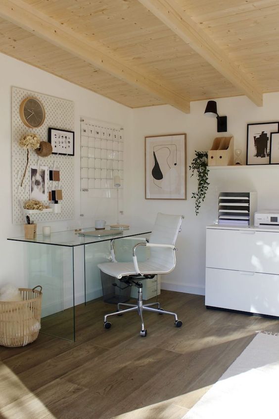 a white shed home office with a glass desk, a white chair, a white storage unit, a ledge with decor, a memo board and some decor